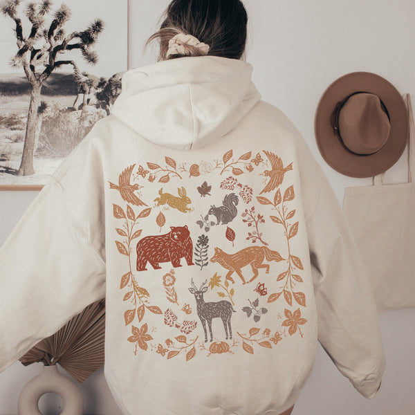This cozy hoodie brings the wonders of the woods to life, with an intricate scene of woodland animals like foxes, bears, and rabbits illustrated on the back, creating a vibrant forest landscape you can carry with you.